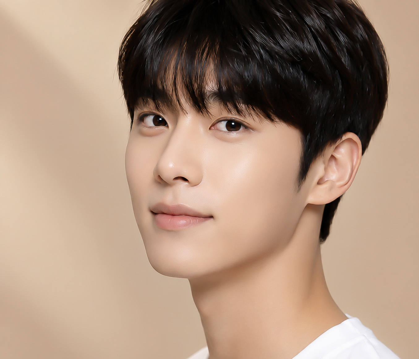 Close-up portrait of a young man with short black hair and a neutral beige background, looking at the camera wearing a white shirt.