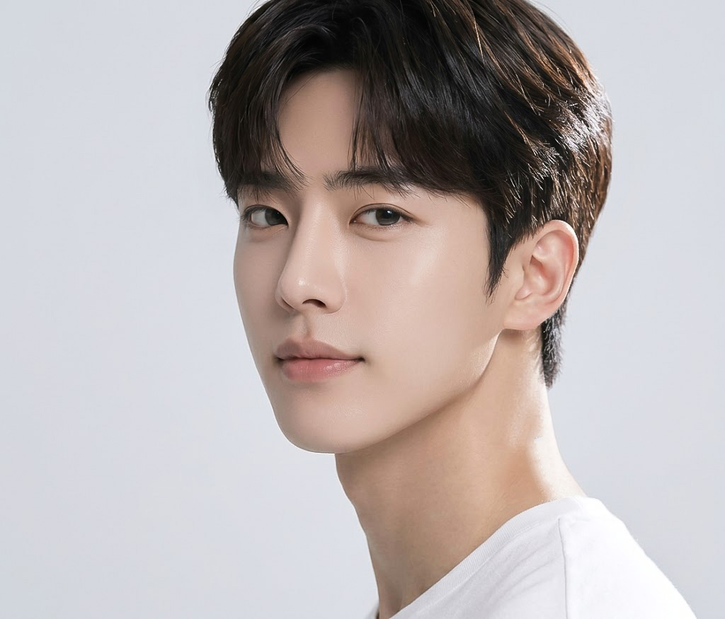 Close-up portrait of a young man with dark hair and a white shirt, looking at the camera against a light background.